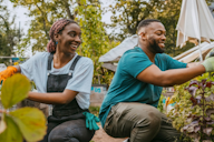 Two people smiling and gardening together in a community garden, wearing gloves and surrounded by plants.